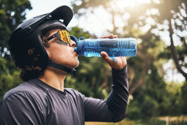 Man, water and bottle in forest for hydration, with cycling equipment and sunglasses by trees for wellness. Cyclist, drink and sports while exercise, workout or training for fitness outside in woods