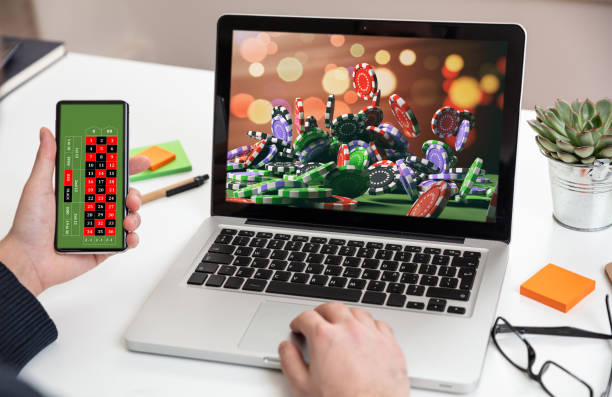 Online gambling, betting concept. Poker chips, casino roulette on laptop and phone screens. Man working with a computer and holding a smartphone, office background